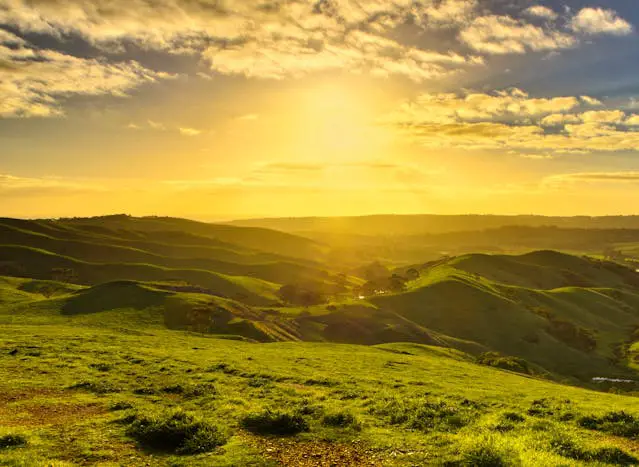 a green hills with a sun shining through the clouds