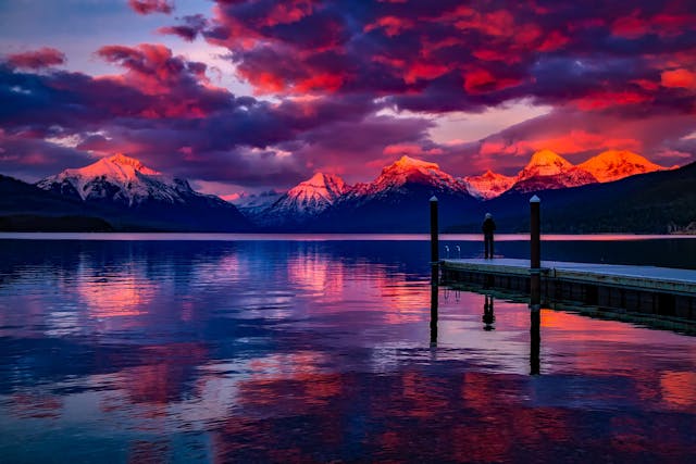 a person standing on a dock on a lake with mountains in the background